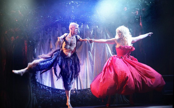 A man and a woman dance. The man is wearing fur-covered trousers and is bare-chested. The woman wears a red shoulder less ball gown.