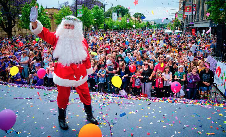 Father Christmas, dressed in his trademark red suit with fur-trimmed sleeves and coat, waves to a crowd of people as he walks along a raised catwalk.