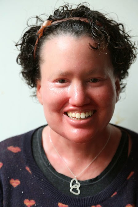 A headshot of Carly smiling . She has short curly black hair and is wearing a black top with red hearts on it and a silver necklace with two hearts on it.