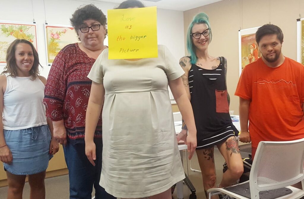 Five people standing smiling at the camera. The person in the centre has a post it note over her face that says love as the bigger picture.