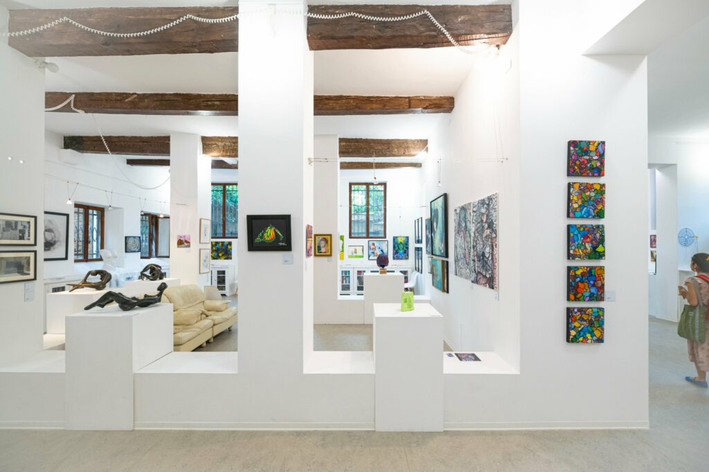  Inside a bright, white art gallery space with variously leveled walls and plinths containing artworks, SKEG's multi-coloured, mosaic-like, small square artworks hang in a vertical straight line to the right. These are surrounded by other sculptures and paintings.