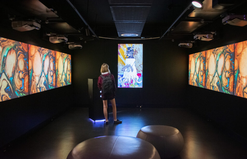 A schoolgirl in a darkly lit room facing a large, portrait screen containing colourful illustrations.