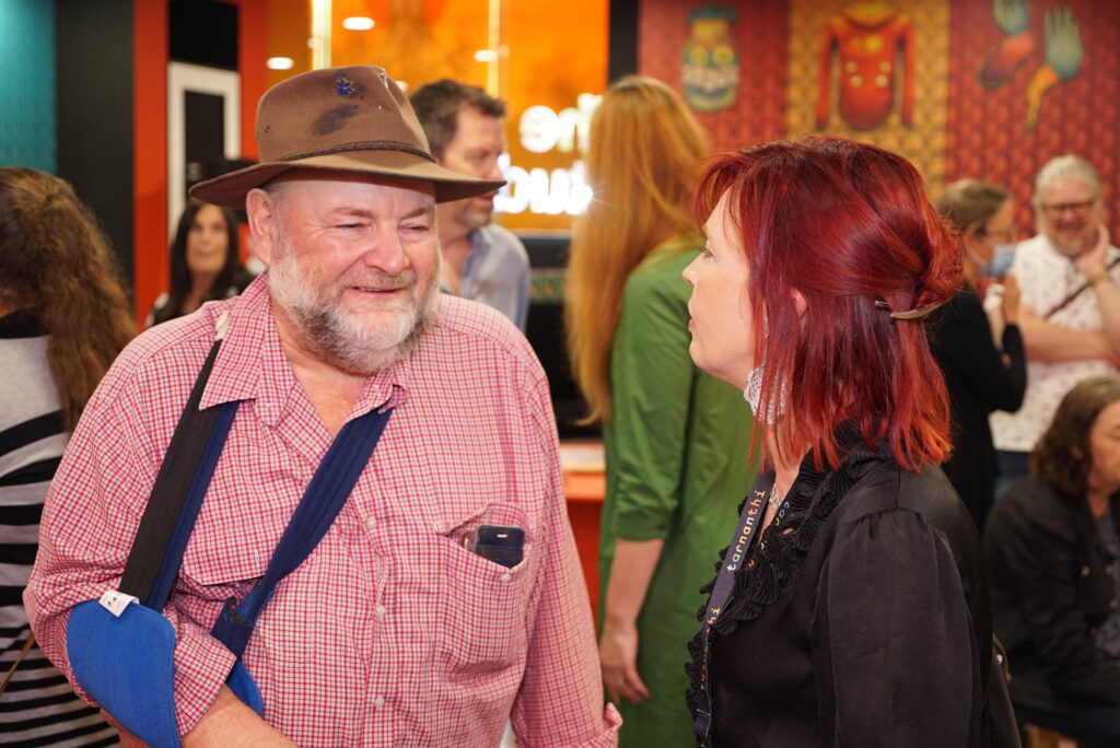 Bundy, an older Caucasian man wearing a pink checkered, long-sleeve shirt, and brown hat, and with his arm in a blue sling, smiles while interacting with a woman with red hair and wearing a black top.