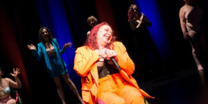 Diana Divine, a performer with red shoulder-length hair and wearing a bright orange two-piece suit, sits in their mobility aid while smiling as they’re surrounded by standing performers who clap at them.