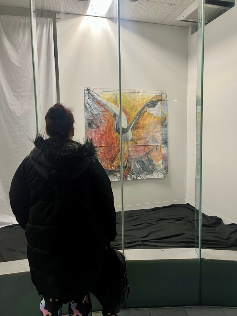 At night, a person in a jacket stands looking at a painting illuminated in a glass window. The orange, red, yellow, white and grey painting depicts a large bird (a jabiru) with outstretched wings.