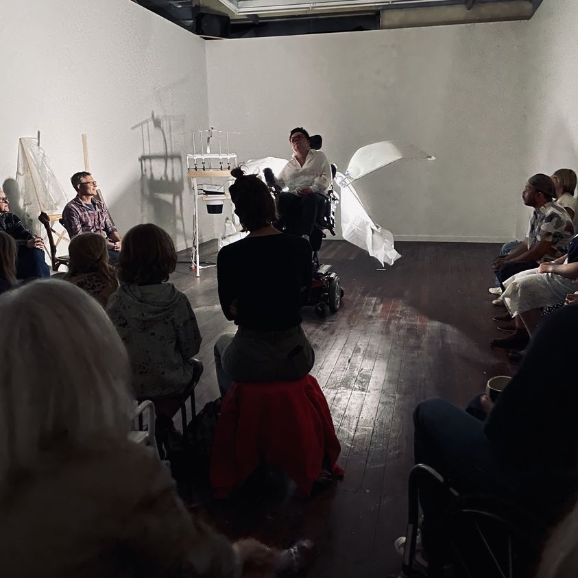 Kirsty, a Caucasian woman, sits in her wheelchair in the centre of a gallery space, the audience looks on attentively, there are white wings on her chair, illuminated by a light behind her.