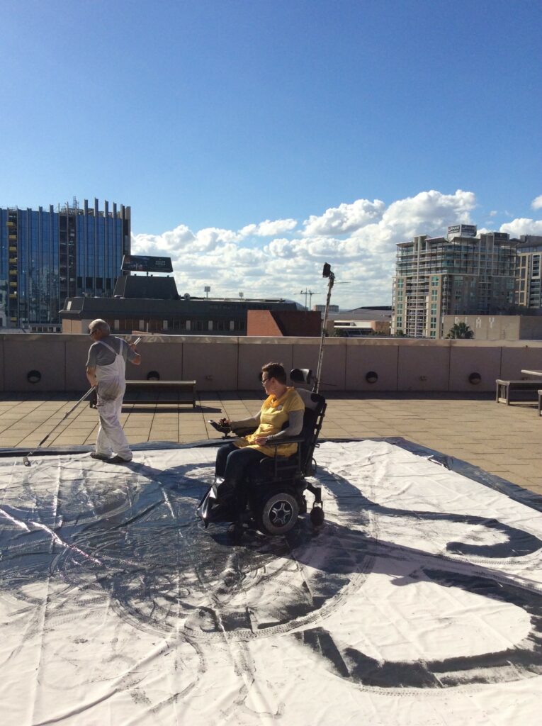 Kirsty Martinsen, a Caucasian woman, in an electronic wheelchair on top of a large cream sheet of material with round black ink patterns. Kirsty is on a large, open rooftop. A blue sky with white clouds and the tops of high-rise buildings are in the background.