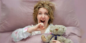 A woman with fluffy short blonde hair holds a hand up to her open mouth as she yawns. She grasps a teddy bear as she lies in a pink bed.